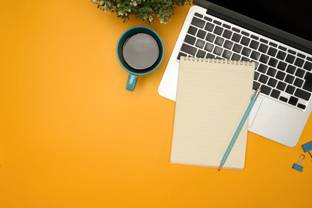 Flat lay of modern desk with a coffee cup, laptop and notebook on yellow background.