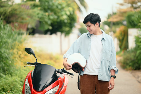 Young asian man holding a helmet standing next to his scooter in a suburban neighborhood.の写真素材