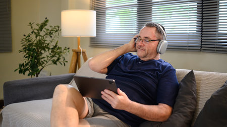 Senior man wearing headphones relaxing on the couch and using a digital tablet.の写真素材