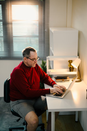 Senior man in red sweater working on a laptop at minimalist desk under desk lamp.の写真素材