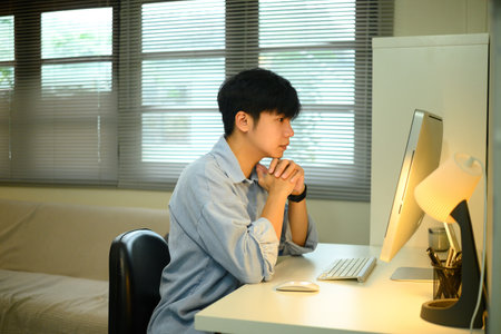 Young man working from home, sitting at desk and concentrating on desktop computer screen.の写真素材