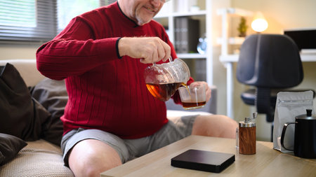Older man in a red sweater pouring coffee from a glass pot into a cup relaxing at home.の写真素材