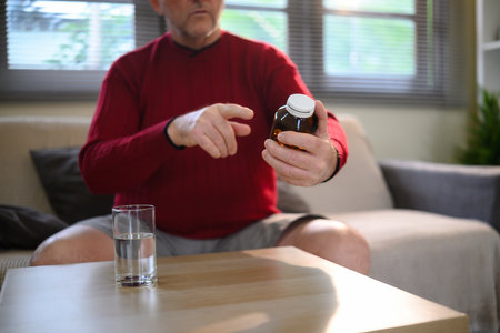 Mature man examining a prescription bottle while seated at home with a glass of water.の写真素材