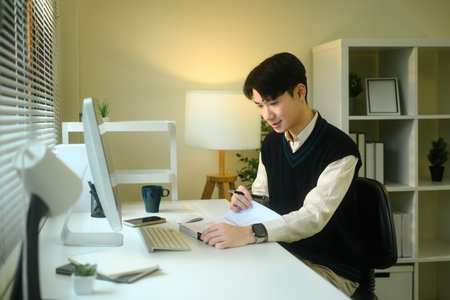 Focused Asian male working on a document while seated at a white desk.の写真素材