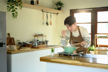 Happy young asian man tasting a freshly prepared meal from a green pot.の写真素材