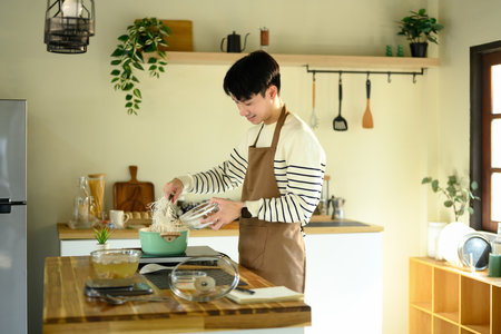 Smiling man in an apron adding noodles to a pot in a sunlit kitchen.の写真素材