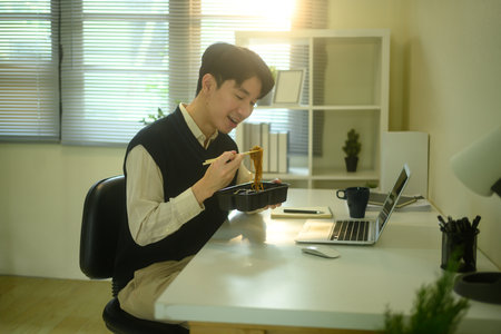 Asian man enjoying a quick meal at his desk in a modern workspace.の写真素材