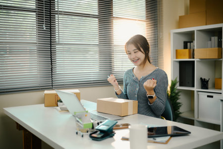Happy young Asian woman celebrating successful sales at desk with laptop and parcel boxes.の写真素材