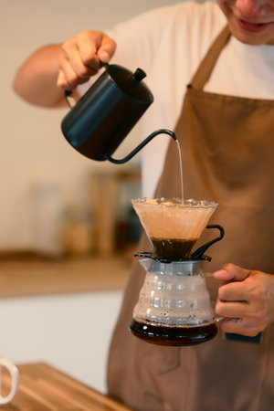 Young man in apron brewing coffee using the pour-over method in warm kitchen.の写真素材