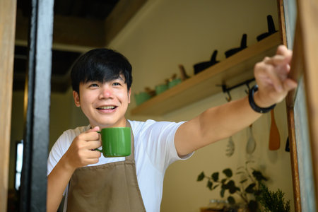 Young man holding a coffee cup smiling warmly as he opens a window.の写真素材