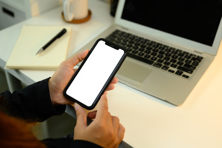 Close up of hands holding smartphone with blank white screen next to laptop.の写真素材