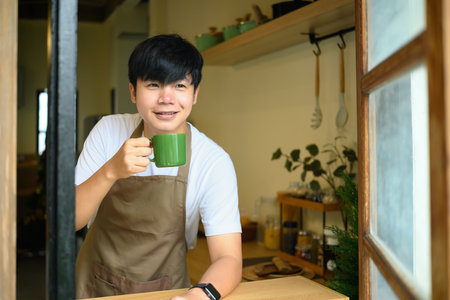 Young man in an apron holding a coffee mug and smiling by the window in a kitchen.の写真素材