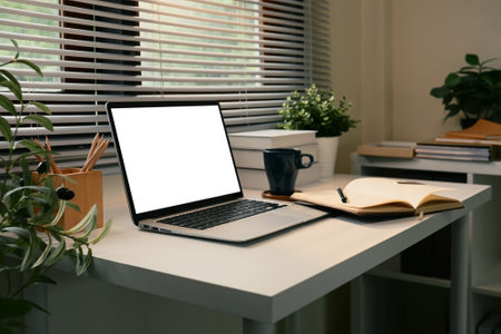 Modern workspace with blank laptop screen, notebook, and coffee mug in a bright room.の写真素材