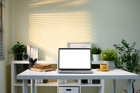 Minimalist desk with laptop, potted plants, alarm clock, and warm toned lighting.の写真素材