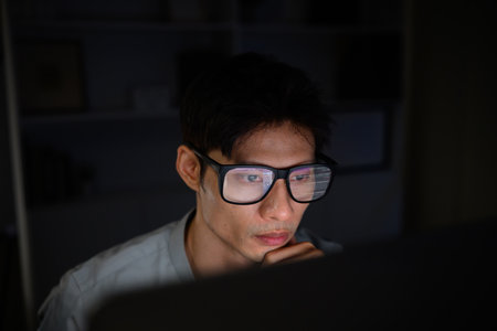 Young man wearing glasses working late at night, focused on computer screen with code reflection.の写真素材