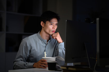Office worker eating instant noodles while working overtime in dimly lit office.の写真素材