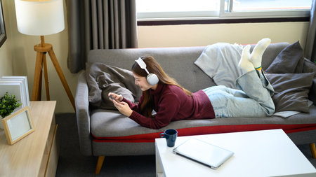Happy woman lounging on sofa with headphones, using mobile phone for entertainment.の写真素材