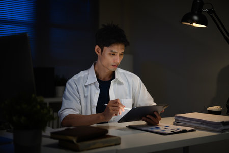 Focused businessman working late at night, analyzing documents on digital tablet in dark office.の写真素材