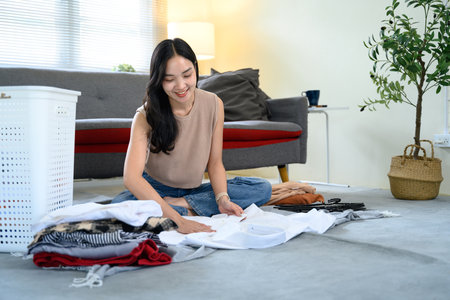 Smiling young woman folding laundry at home, organizing clothes neatly on the floor.の写真素材