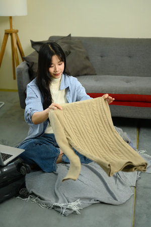 Young woman folding a beige knitted sweater while packing clothes for a trip at home.の写真素材