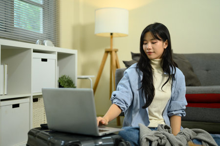 Young woman sitting on the floor with a suitcase, using a laptop to plan and organize her trip.の写真素材