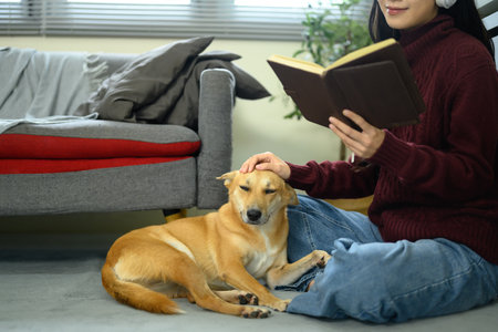 Woman sitting on the floor reading a book while gently petting her dog in a living room.の写真素材