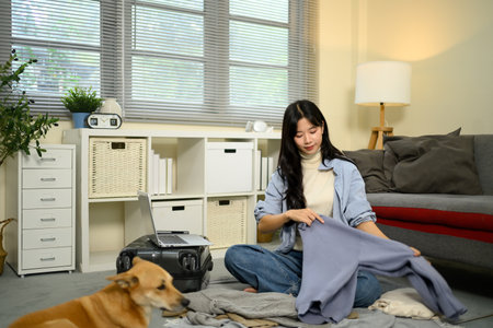 Young woman packing clothes into suitcase while preparing for travel, with dog lying nearby.