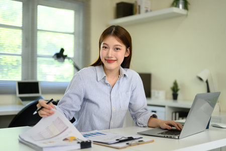 Smiling young businesswoman working on laptop while reviewing financial reports.の写真素材