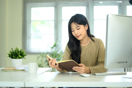 Young woman reading a notebook while sitting at desk with computer and coffee cup.の写真素材
