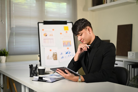 Businessman in black suit working on digital tablet with financial charts in background.の写真素材