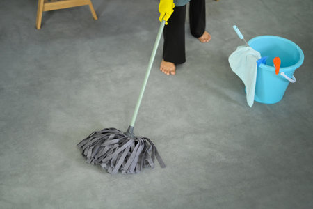 Person wearing yellow gloves mopping a gray floor with blue bucket and cleaning tools.