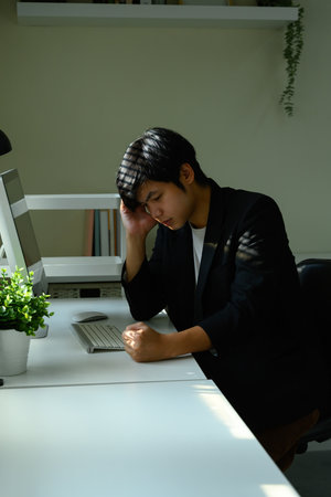 Young businessman at desk with hand on head, appearing tired and overwhelmed by workload.