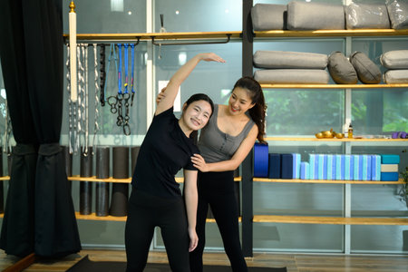 Female trainer assisting young woman with side stretching exercise in a modern yoga studio.