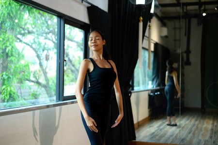 Graceful ballerina standing near window with eyes closed in elegant ballet pose.