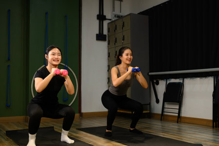 Female trainer and student working out together, holding weights and doing squats for muscle...