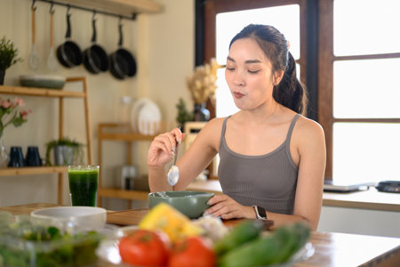 Smiling woman in activewear eating yogurt topped with fresh berries in a bright kitchen.の写真素材