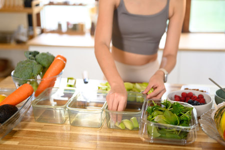 Woman organizing sliced fruits and fresh vegetables into meal containers on a wooden kitchen...