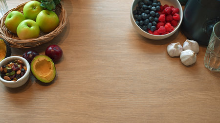 Flat lay of healthy fresh fruits, avocado, nuts, and berries arranged around wooden table with copy...