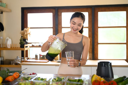Smiling woman pouring freshly blended green smoothie into a glass in a bright modern kitchen.の写真素材