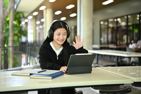 Smiling Gen Z student wearing headphones making a video call on tablet during online learning...