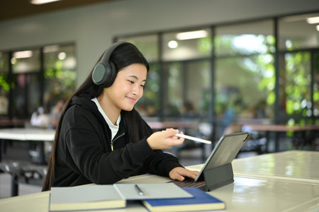 Gen Z student using tablet and stylus with headphones, smiling while studying in a modern open school environment..の写真素材