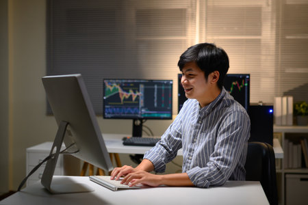 Young Asian trader analyzing financial charts on computer at night, focused on investment strategy.の写真素材