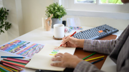 Closeup creative woman sketching logo ideas in notebook with color samples and pencils in bright modern workspace.の写真素材