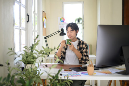 Creative woman relaxing with coffee at her desk during break in bright modern workspace with computer and design tools.の写真素材