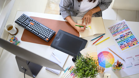 Top view creative woman sketching logo ideas in notebook with color samples and pencils.