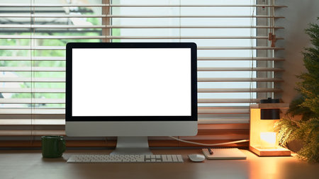 Minimalist workspace with computer, desk lamp, and coffee mug by window in warm light..の写真素材