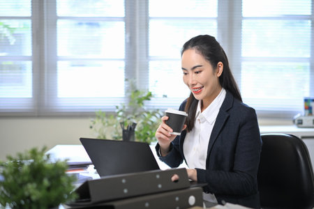 Smiling businesswoman drinking coffee while working on laptop in bright office, representing balance between productivity and relaxation.の写真素材