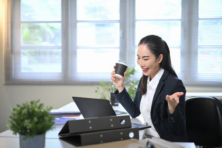 Businesswoman holding coffee cup while consulting online with client via laptop, representing digital communication and remote teamwork.の写真素材