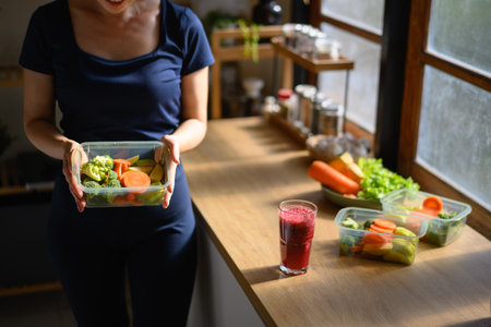 A woman holding a food container with fresh vegetables and a smoothie on the table, symbolizing a healthy and mindful lifestyle at home.の写真素材