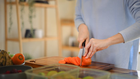 Closeup of young female hands chopping fresh orange carrot on board while in modern kitchen.の写真素材